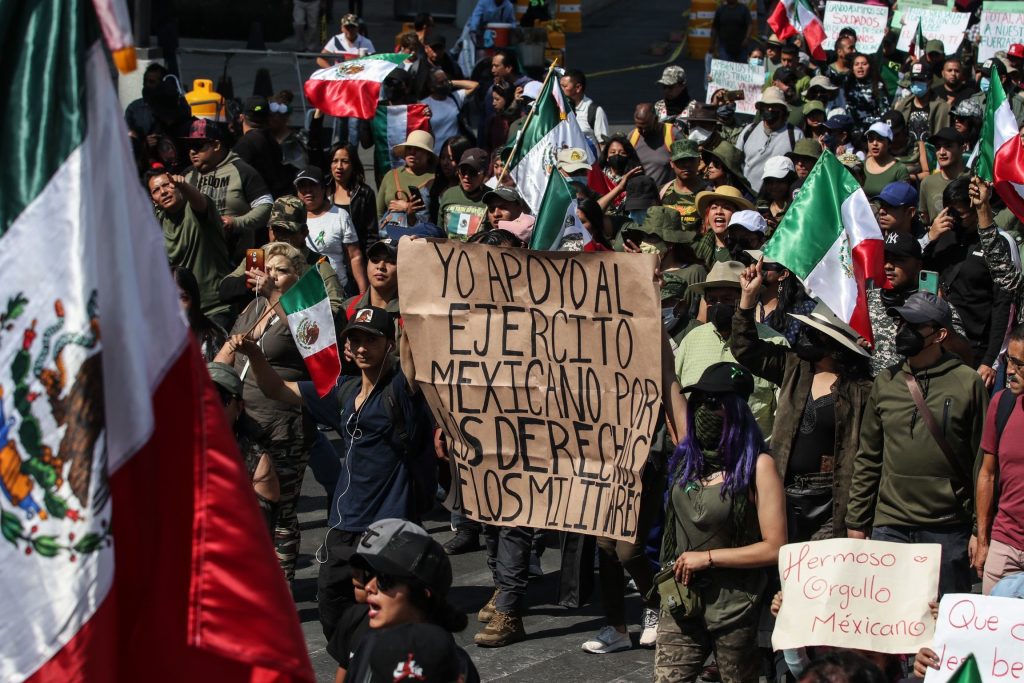 At least 40 people in an urban setting. Many wear dark green and are waving Mexican flags or carrying hand-written signs.