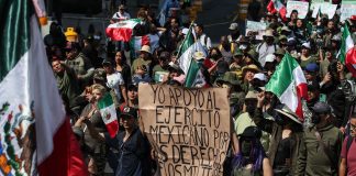 At least 40 people in an urban setting. Many wear dark green and are waving Mexican flags or carrying hand-written signs.