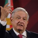 A grey-haired man wearing a suit and tie gestures with the red, white and green of the Mexican flag visible in the background.