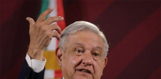 A grey-haired man wearing a suit and tie gestures with the red, white and green of the Mexican flag visible in the background.