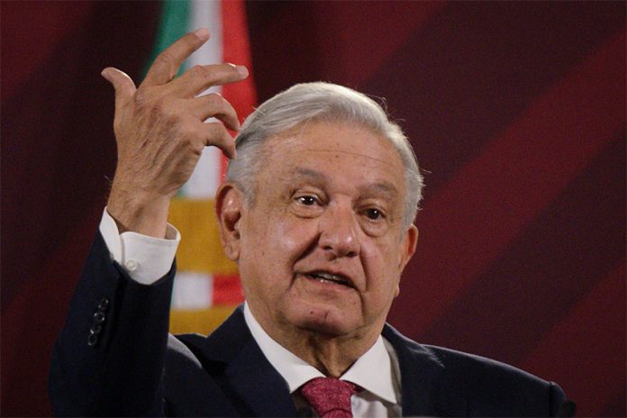 A grey-haired man wearing a suit and tie gestures with the red, white and green of the Mexican flag visible in the background.