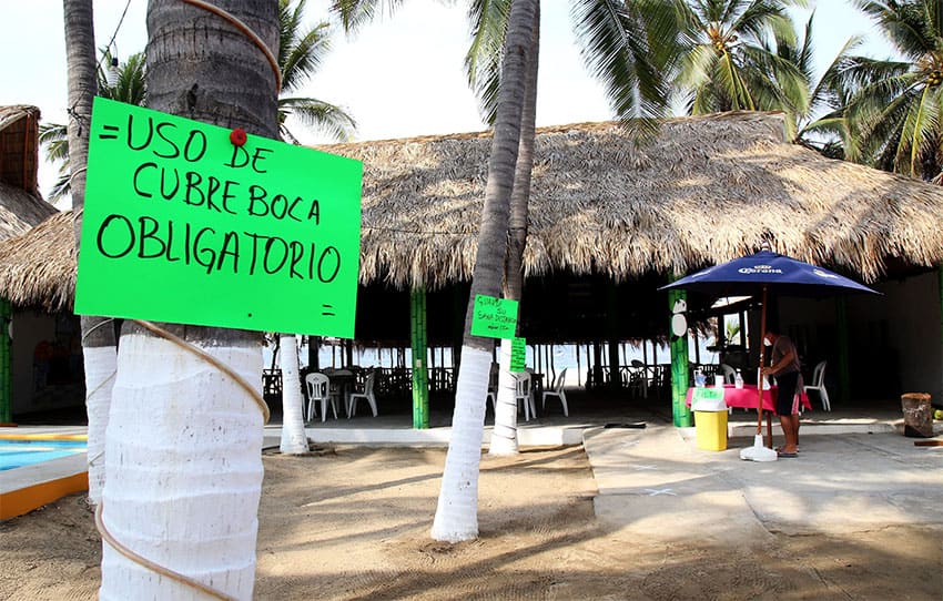A hand written sign saying "uso de cubreboca obligatorio" pinned to the truck of a palm tree with a palapa in the background.