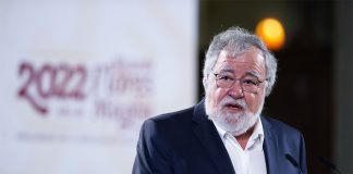 A grey-bearded, light-skinned man with glasses speaks at a podium.