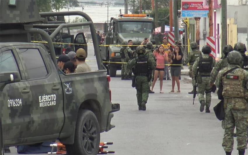 Soldiers in uniform walk near several camo vehicles in a cordoned-off area of the street, with residents in everyday clothing at the edges of the area.