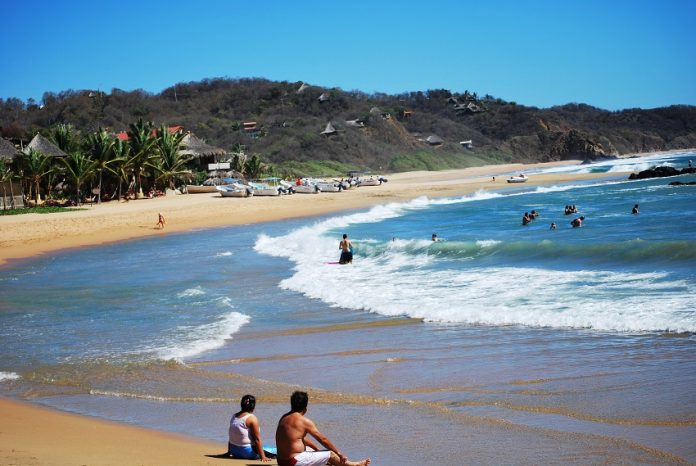 Two people sit on the beach in San Agustinillo, Oaxaca as others bathe in the surf.