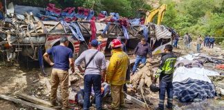 Rescue crew stands in front of totaled bus, bodies covered in sheets are visible.