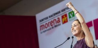 Claudia Sheinbaum stands at a podium in front of a sign showing the logos of Morena and its allies