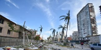 Pieces of debris litter a street lined with damaged palm trees and hotels.