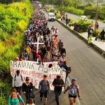 A crowd of people walk down a road carrying a cross and a banner that says "Caravan Migrante."