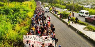 A crowd of people walk down a road carrying a cross and a banner that says "Caravan Migrante."