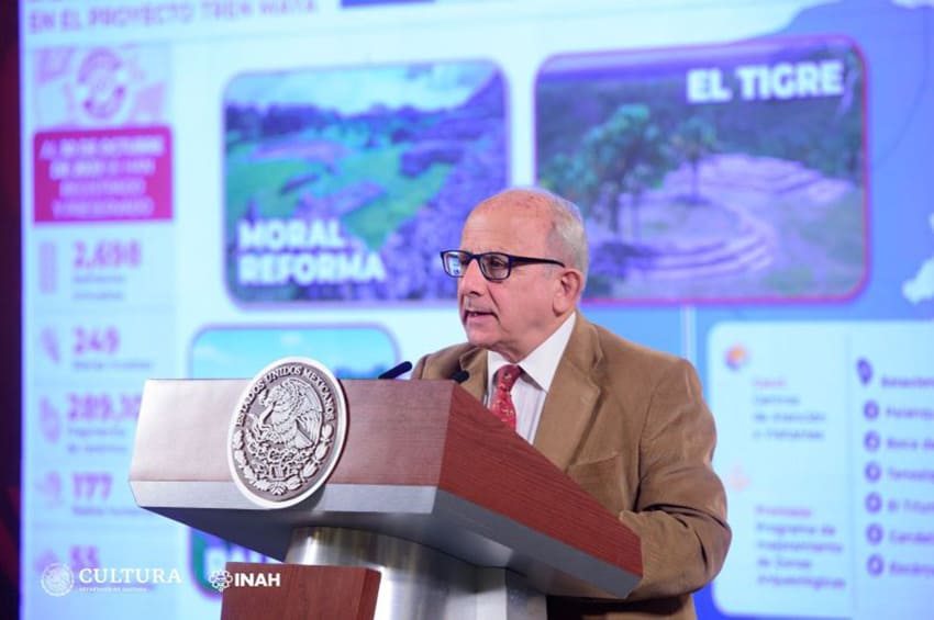 An older man with glasses speaks at a podium in front of a screen.