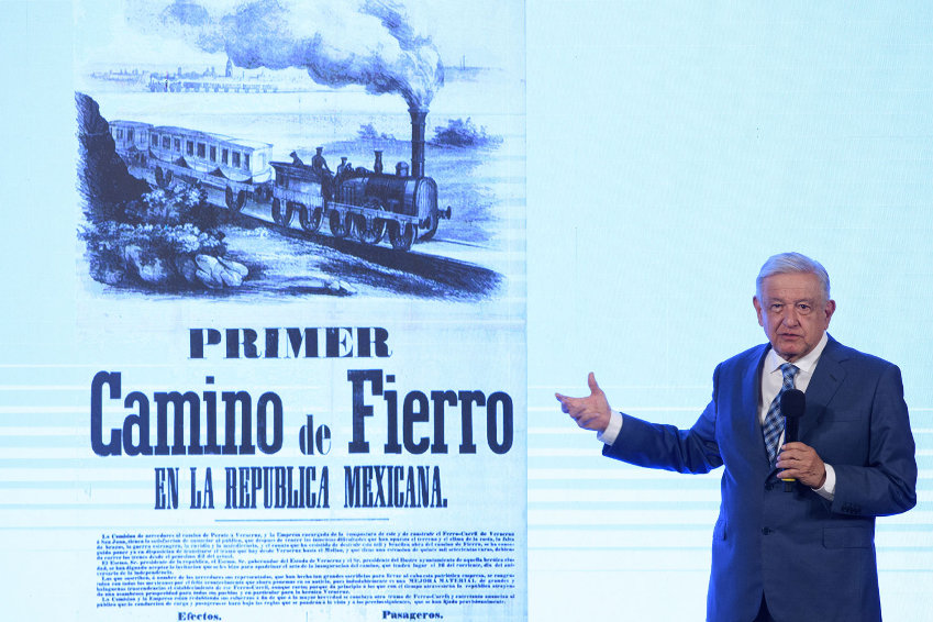 El presidente López Obrador señala una pantalla detrás de él, mostrando una ilustración de un tren histórico.