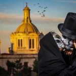 A fancily dressed man and women with their face painted as skulls kiss in front of a cathedral.