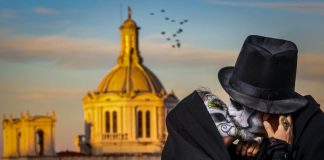 A fancily dressed man and women with their face painted as skulls kiss in front of a cathedral.
