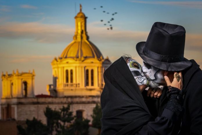 A fancily dressed man and women with their face painted as skulls kiss in front of a cathedral.