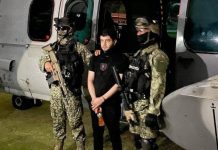 El Nini, a man in his 30s or 40s, stands between two Mexican soldiers in front of a helicopter.