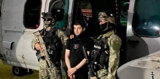 El Nini, a man in his 30s or 40s, stands between two Mexican soldiers in front of a helicopter.