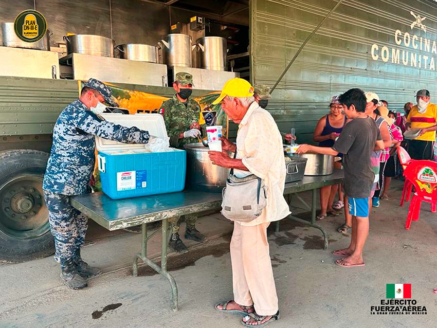 Military members in camo uniforms serve food to a line of people of various ages.