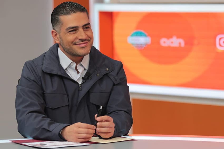 A man in a black coat smiles on a TV news set