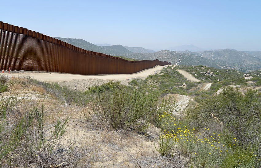 The Mexico-US border wall runs through the desert with mountains behind it.
