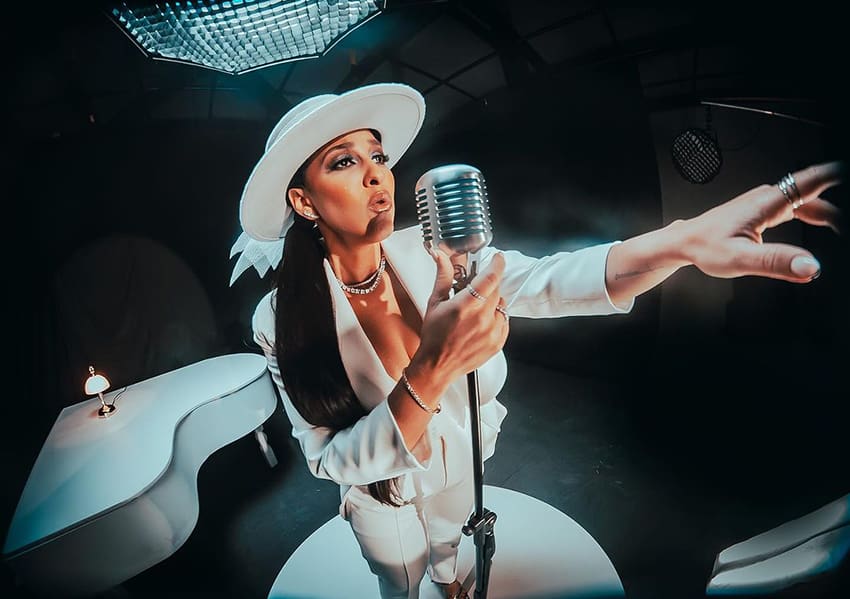 A fisheye photo of a woman in a white hat and dramatic makeup singing with a microphone