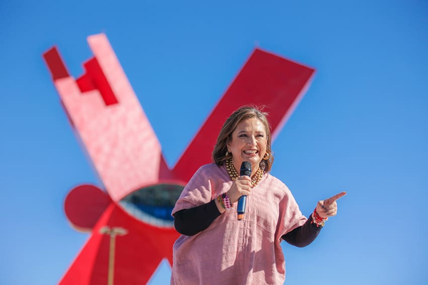 Xóchitl Gálvez habla por un micrófono con un monumento rojo abstracto al fondo