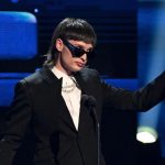 Singer Peso Pluma on stage with a mullet, a black jacket and dark glasses.