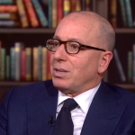 A bald man with glasses and a suit sitting down in an office or library
