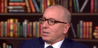 A bald man with glasses and a suit sitting down in an office or library
