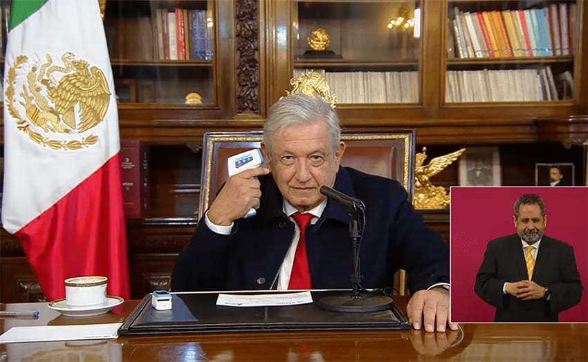AMLO in a suit in his office, presses a thermometer to his head and looks into the camera