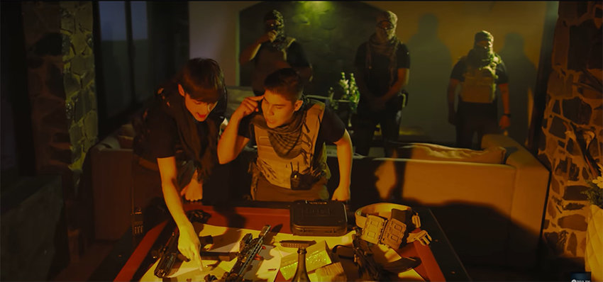 Two men point at a map on a table with guns in a dimly lit room