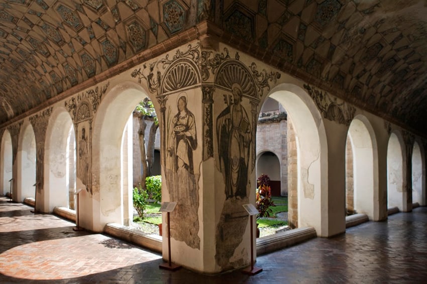 El patio de un convento con jardines apenas visibles al otro lado de arcos elaboradamente pintados.