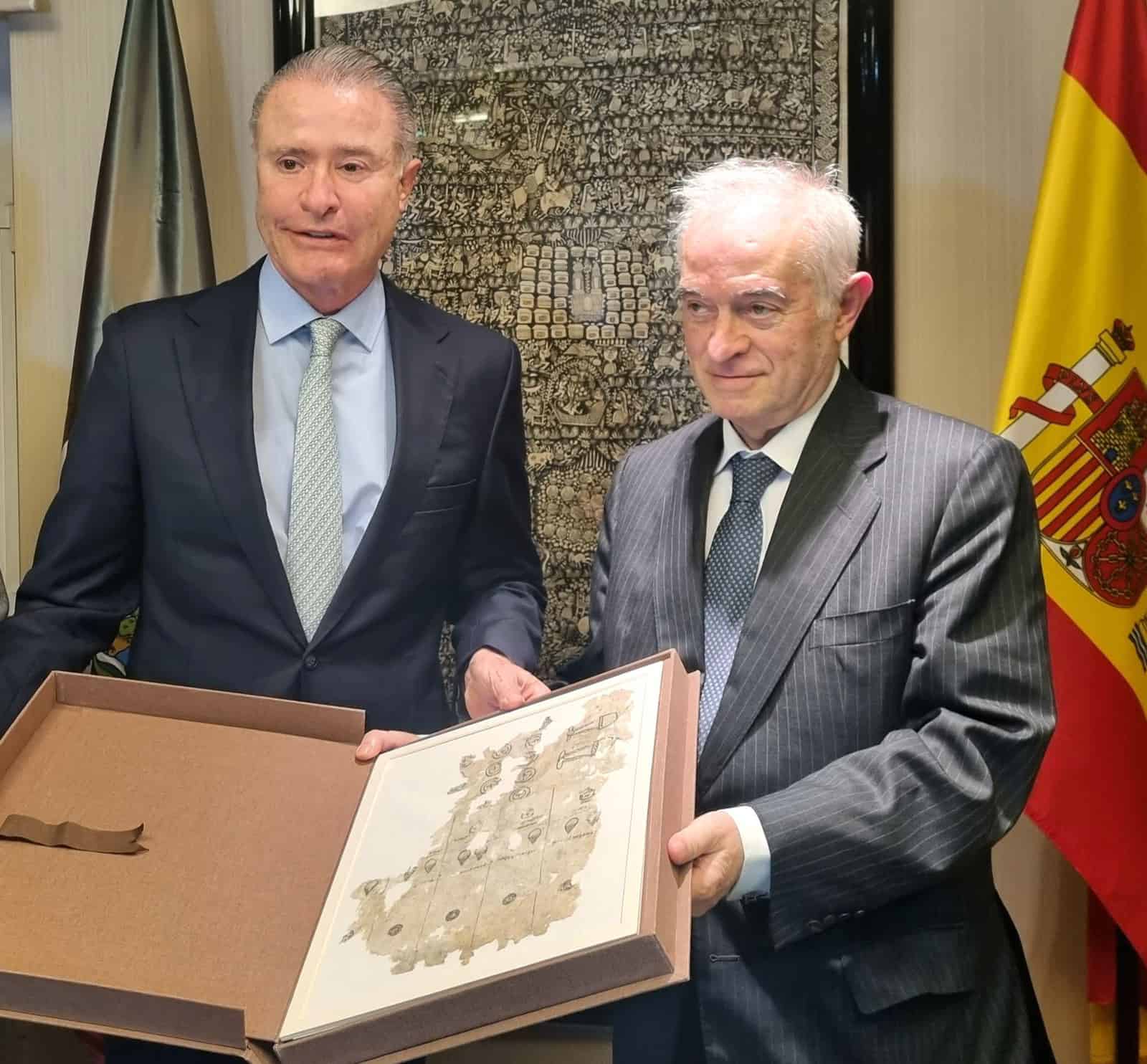 Two men in suits hold a binder displaying an old document, standing in front of a Spanish flag.