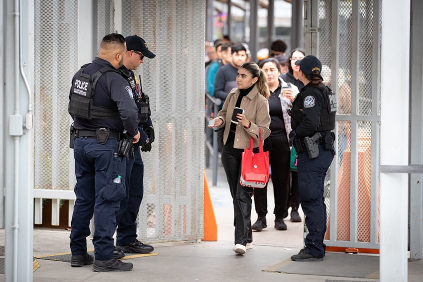 A line of people wait at a metal gate manned by CBP guards