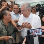 President López Obrador, dressed in a white guayabera, shakes the hand of a supporter while others crowd around and take pictures.