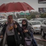 Two women share a red umbrella in the rainy next to a busy road