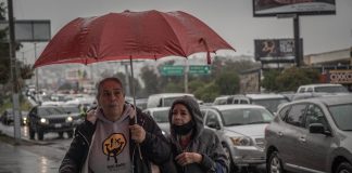 Two women share a red umbrella in the rainy next to a busy road