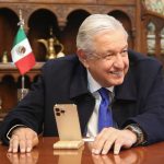 López Obrador sits in front of a smartphone on a phone stand, smiling