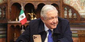 López Obrador sits in front of a smartphone on a phone stand, smiling