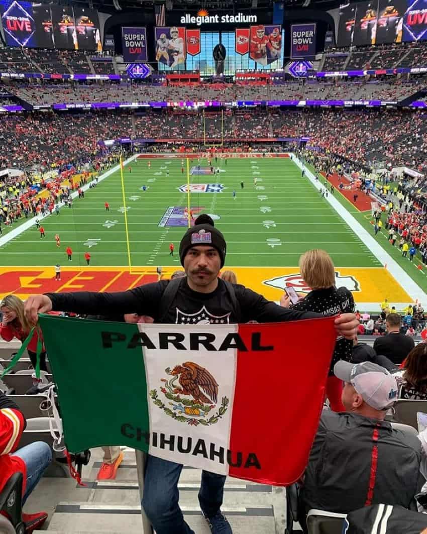 Un hombre sostiene una bandera mexicana leyendo "Parral, Chihuahua," mientras en las gradas del Super Bowl