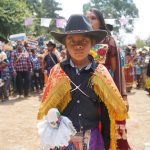 A child wearing a cowboy hat and traditional clothing looks at the camera as festival dancers prepare to perform in the background.