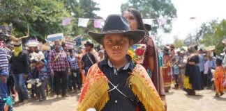 A child wearing a cowboy hat and traditional clothing looks at the camera as festival dancers prepare to perform in the background.