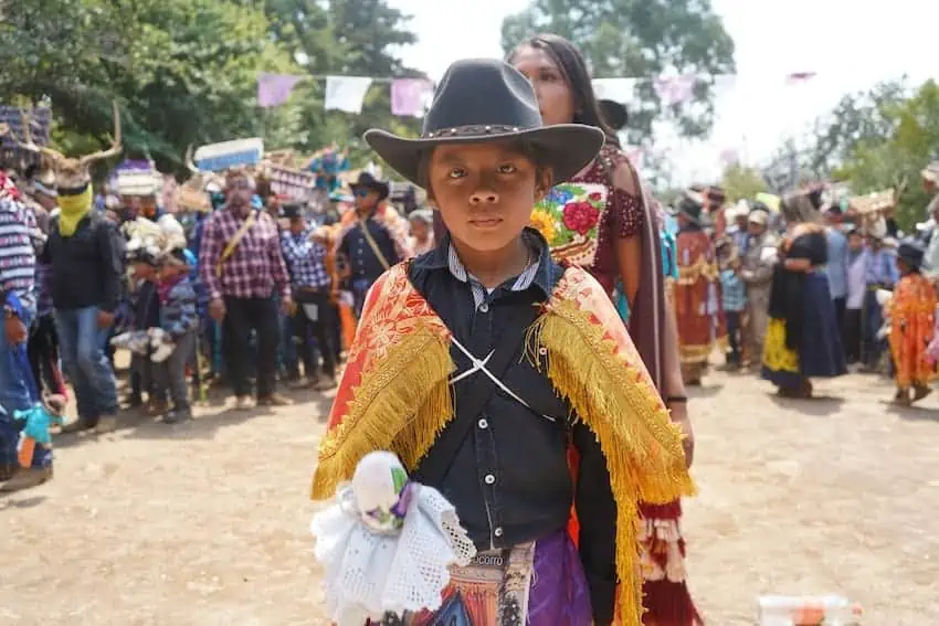 A child wearing a cowboy hat and traditional clothing looks at the camera as festival dancers prepare to perform in the background.