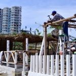 A builder hammers a wooden palapa post in Acapulco, with a multi-story building in the background.
