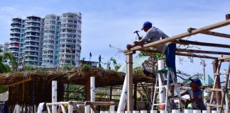 A builder hammers a wooden palapa post in Acapulco, with a multi-story building in the background.