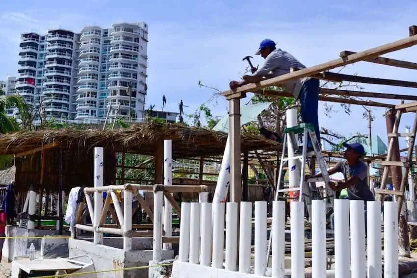 A builder hammers a wooden palapa post in Acapulco, with a multi-story building in the background.