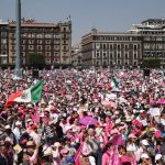 A crowd of people wearing pink and white and waving a Mexican flag fill a plaza, with Mexico's National Palace in the background