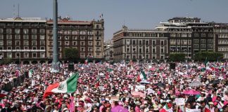A crowd of people wearing pink and white and waving a Mexican flag fill a plaza, with Mexico's National Palace in the background