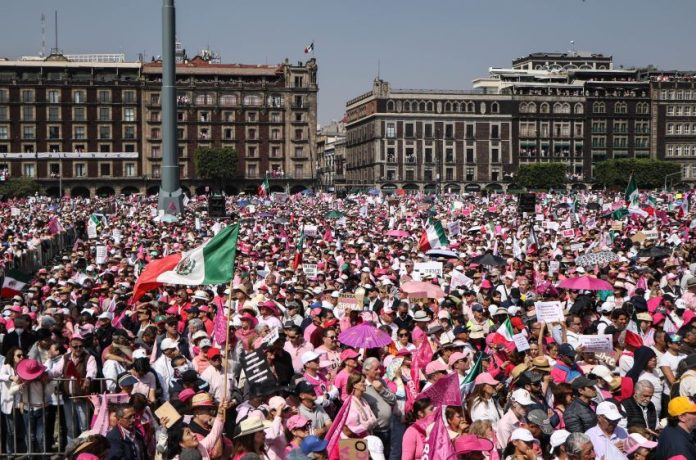 A crowd of people wearing pink and white and waving a Mexican flag fill a plaza, with Mexico's National Palace in the background