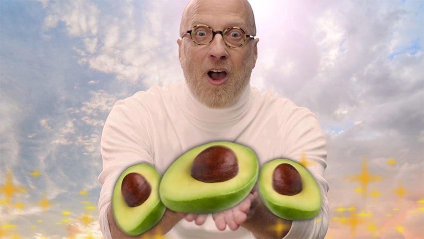 A bald white man in in a pink turtleneck offers large, bright green avocados toward the camera